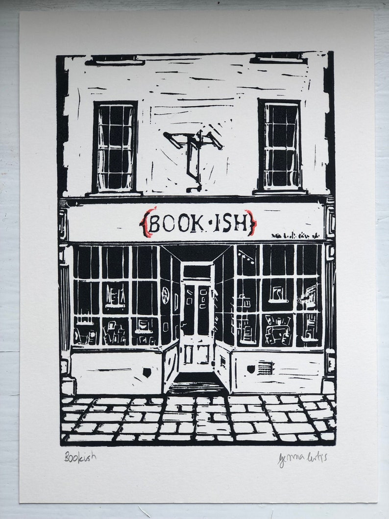 May include: Black and white linocut print of a shop front with the text "BOOK.ISH" above the door. The shop front has two windows and a door with a sign above the door. The print is signed "Bookish" and "Gemma Curtis" in the bottom corners.