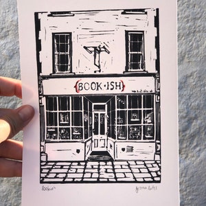May include: Black and white linocut print of a shop front with the text "BOOK.ISH" above the door. The print is signed "Bookish" and "Gemma Cutts" in the bottom corners.