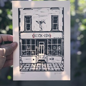 May include: Black and white linocut print of a shop front with the text "BOOK-ISH" above the door. The shop front has two windows and a door with a sign above the door that says "BOOK-ISH". The print is signed "Bookish" and "Gemma Curtis" in the bottom corners.