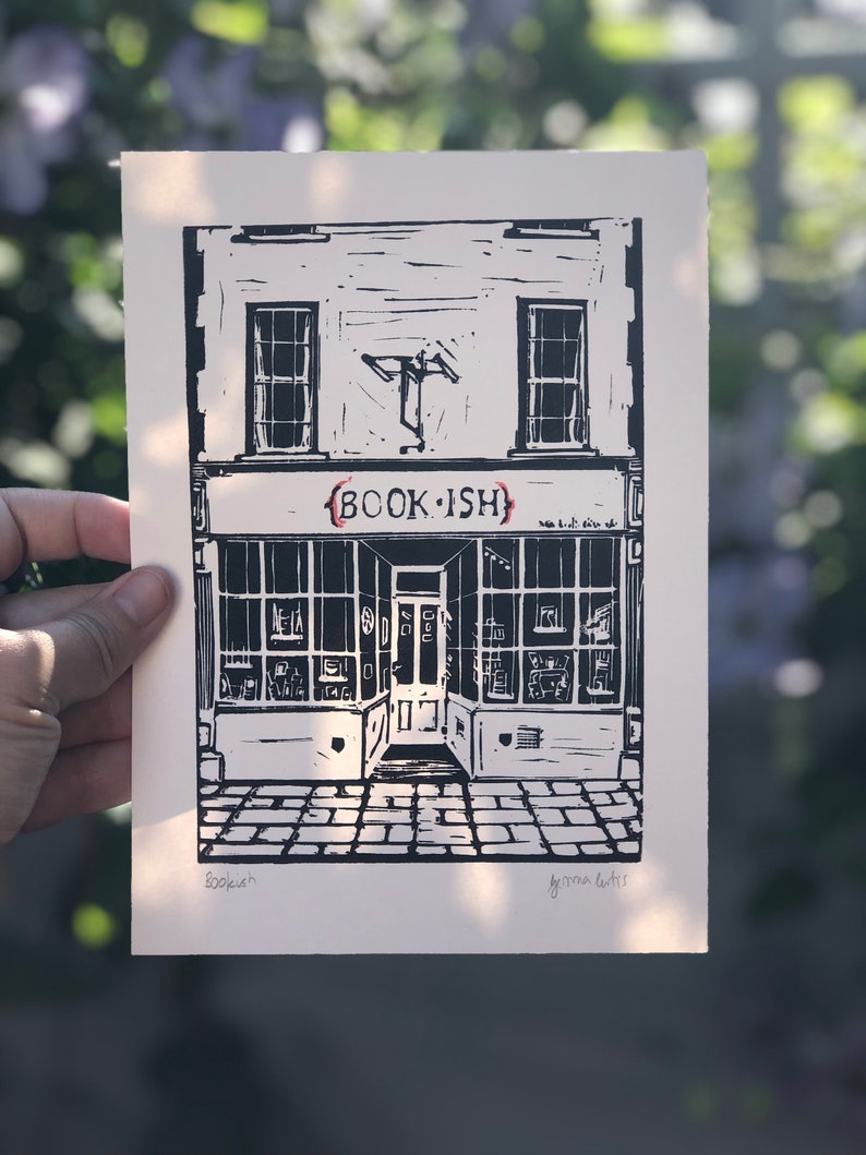 May include: Black and white linocut print of a shop front with the text "BOOK-ISH" above the door. The shop front has two windows and a door with a sign above the door that says "BOOK-ISH". The print is signed "Bookish" and "Gemma Curtis" in the bottom corners.
