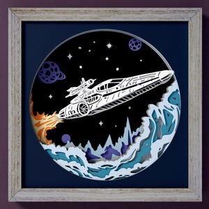 Puede incluir: Una obra de arte en papel en capas enmarcada que representa una nave espacial blanca en vuelo contra un fondo espacial negro con planetas y estrellas. La nave espacial tiene llamas naranjas. El marco es gris claro. El texto "SVG", "DXF", "EPS" y "PDF" está a la izquierda.