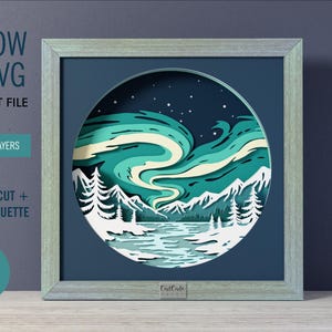 Op de afbeelding: Een 23x23 cm shadow box SVG digitaal snijbestand met 12 genummerde lagen. Het cirkelvormige ontwerp toont een winterlandschap met witte bergen, groenblijvende bomen, een bevroren meer en een levendig groenblauw en crèmekleurig noorderlicht.