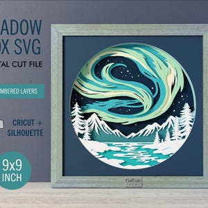 May include: A 9x9 inch shadow box SVG digital cut file featuring 11 numbered layers. The artwork depicts a circular scene with a green and blue aurora borealis above white snow-covered mountains, pine trees, and a river, compatible with Cricut and Silhouette.