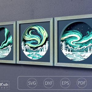 May include: Three square framed wall art pieces featuring layered paper cutouts of the aurora borealis over snowy mountains and pine trees. The artwork displays shades of teal, green, and white against a dark blue sky. Text reads CutCute paper.
