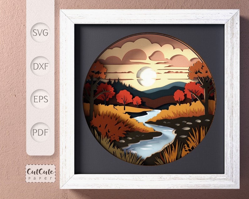 May include: A paper cut art piece depicting a landscape with a river winding through a forest. The scene is set in autumn with colorful trees and a bright blue sky. The art is framed in a white frame.