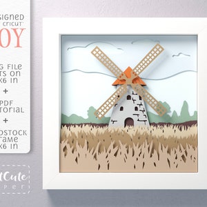 May include: A white frame with a papercraft windmill scene inside. The windmill is made of brown, white, and orange paper. The scene includes a field of brown grass and a blue sky with white clouds. The frame is 6x6 inches. The text "CutCute Paperi" is in the bottom left corner.