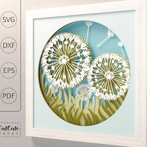 May include: White framed paper art featuring a layered design of dandelions. The dandelions are white with intricate cut details, set against a blue and green background. The artwork is in a square frame, with the text "SVG", "DXF", "EPS", and "PDF" on the left.