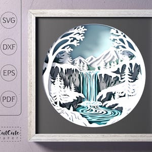 Op de afbeelding: Een wit papieren snijkunstwerk van een waterval in een besneeuwd berglandschap. Het ontwerp is ingelijst in een wit frame met een grijze passe-partout.