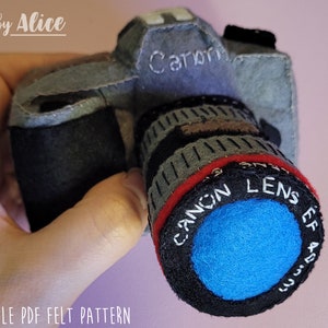 May include: A grey felt camera with a black lens and the word "Canon" embroidered on the front. The lens has a red and black ring with the words "Canon Lens EF 45-250mm" embroidered on it.