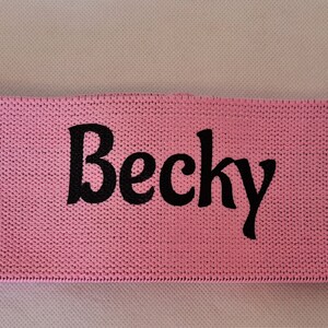 Puede incluir: Banda de resistencia de tela rosa con el nombre "Becky" impreso en negro.