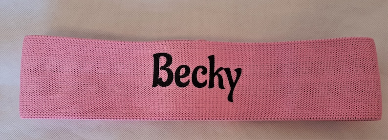 Puede incluir: Banda de resistencia de tela rosa con el nombre "Becky" impreso en negro.