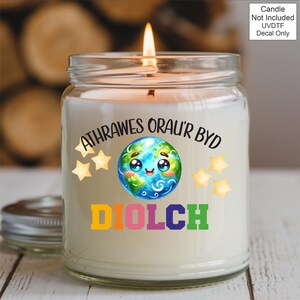 May include: A lit candle in a glass jar with a silver lid. The jar has a decal with a smiling Earth graphic, stars, and the words "ATHRAWES ORAU'R BYD" and "DIOLCH" in colorful letters. The background is blurred.