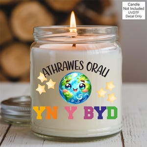May include: A lit candle in a glass jar with a silver lid. The jar has a decal with a cartoon Earth, stars, and text in multiple colors. The text reads "ATHRAWES ORAU" and "YN Y BYD". The background is blurred.