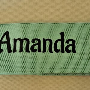 Puede incluir: Una banda de resistencia de tela verde claro con el nombre "Amanda" bordado en negro.