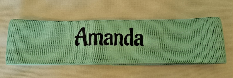 Puede incluir: Una banda de resistencia de tela verde claro con el nombre "Amanda" bordado en negro.
