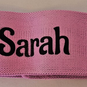 Puede incluir: Banda de resistencia de tela rosa con el nombre "Sarah" bordado en negro.