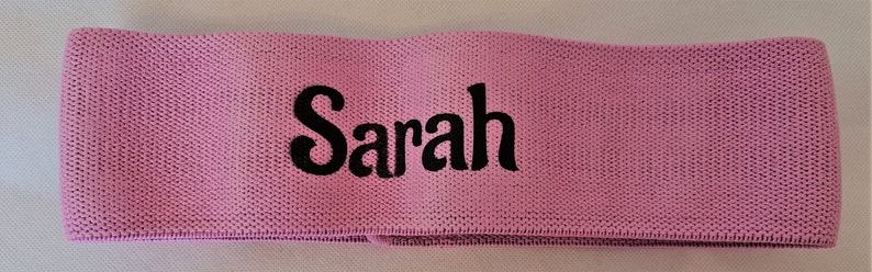 Puede incluir: Banda de resistencia de tela rosa con el nombre "Sarah" bordado en negro.