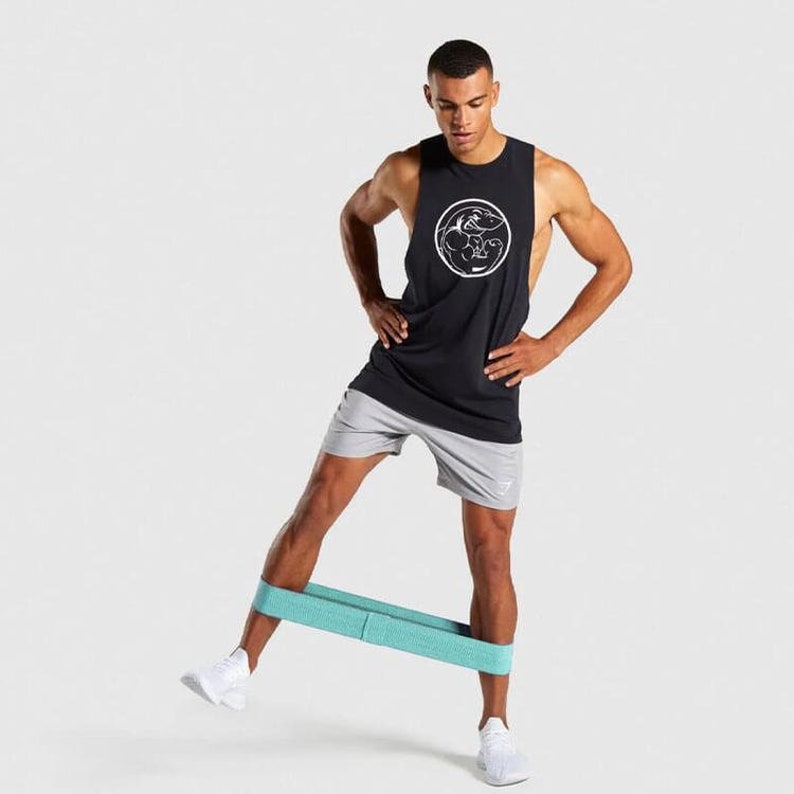 Puede incluir: Una persona que lleva una camiseta sin mangas negra con un logotipo blanco y pantalones cortos grises, utilizando una banda de resistencia turquesa para un ejercicio de piernas.