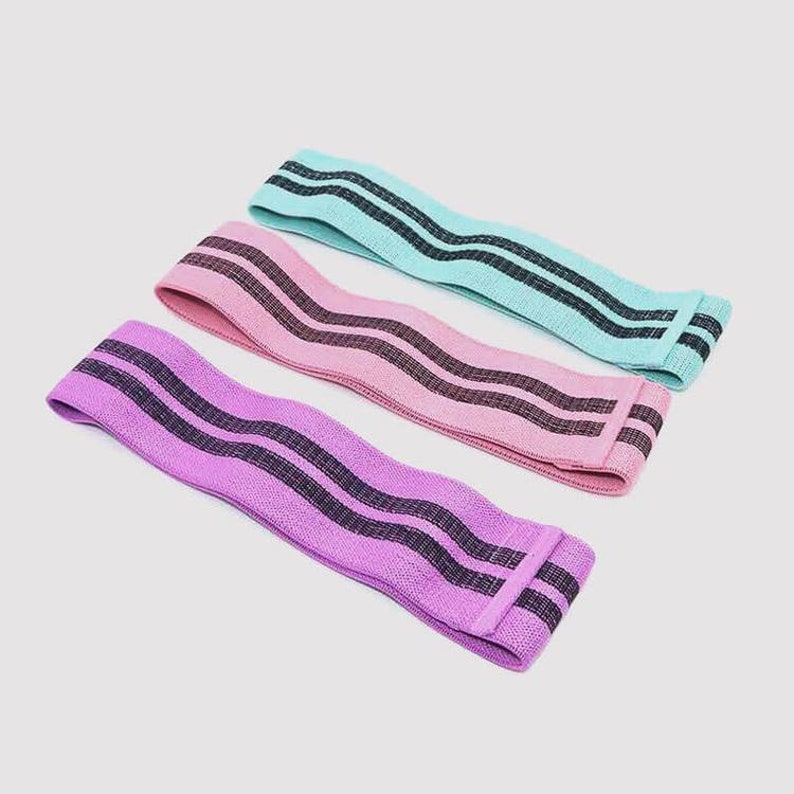 Puede incluir: Tres bandas de resistencia de tela en diferentes colores: morado claro, rosa y azul claro. Cada banda tiene dos rayas negras que recorren toda la longitud de la banda.