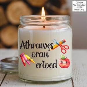 May include: A lit candle in a glass jar with a silver lid. The candle has a decal with the words "Athrawes orau erioed" and illustrations of crayons, scissors, a pencil, and a smiling apple. The text "Candle Not Included UVDTF Decal Only" is also visible.