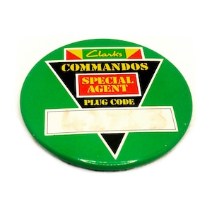 May include: A green round button with a black triangle in the centre. The triangle has the words "Commandos Special Agent Plug Code" in yellow and red lettering. The button also has the word "Clarks" in yellow lettering.