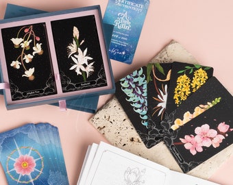 Puede incluir: Una baraja de cartas de oráculo A Walk in the Rain con un libro guía. Las cartas presentan ilustraciones de flores y plantas sobre un fondo negro. El libro guía tiene un dibujo lineal de una flor de cerezo en la portada.