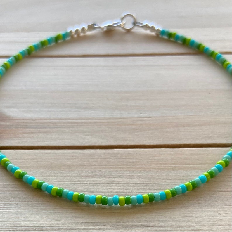 Puede incluir: Un delicado collar de cuentas con cuentas verdes y azules alternadas. El collar tiene un cierre plateado.