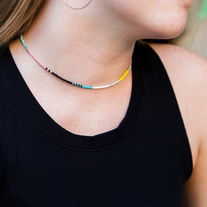 May include: A close-up of a woman wearing a black tank top with a colorful beaded necklace. The necklace features pink, black, turquoise, white, and yellow beads.