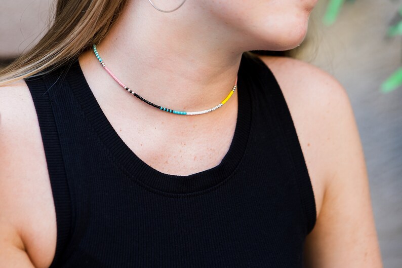 May include: A close-up of a woman wearing a black tank top with a colorful beaded necklace. The necklace features pink, black, turquoise, white, and yellow beads.