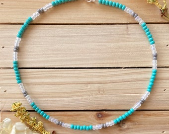 May include: A beaded necklace featuring turquoise, clear, and gray beads. The necklace is strung on a silver-colored chain and clasp. The beads are small and evenly spaced, creating a delicate and colorful accessory.