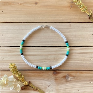 May include: A white beaded necklace with black, green, and blue beads. The necklace is on a wooden surface.