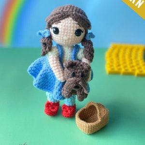 May include: Crochet pattern for a Dorothy doll from the Wizard of Oz. The doll is holding a brown dog and has a blue dress, red shoes, and brown hair. The pattern is available as a PDF download.