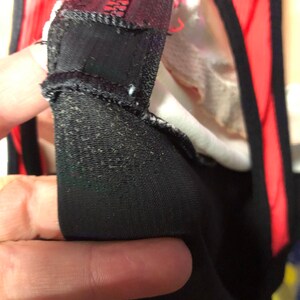 May include: Close-up of black fabric with a textured surface, possibly part of a garment. The fabric is folded over, revealing a red and white interior. The image highlights the stitching and construction details.