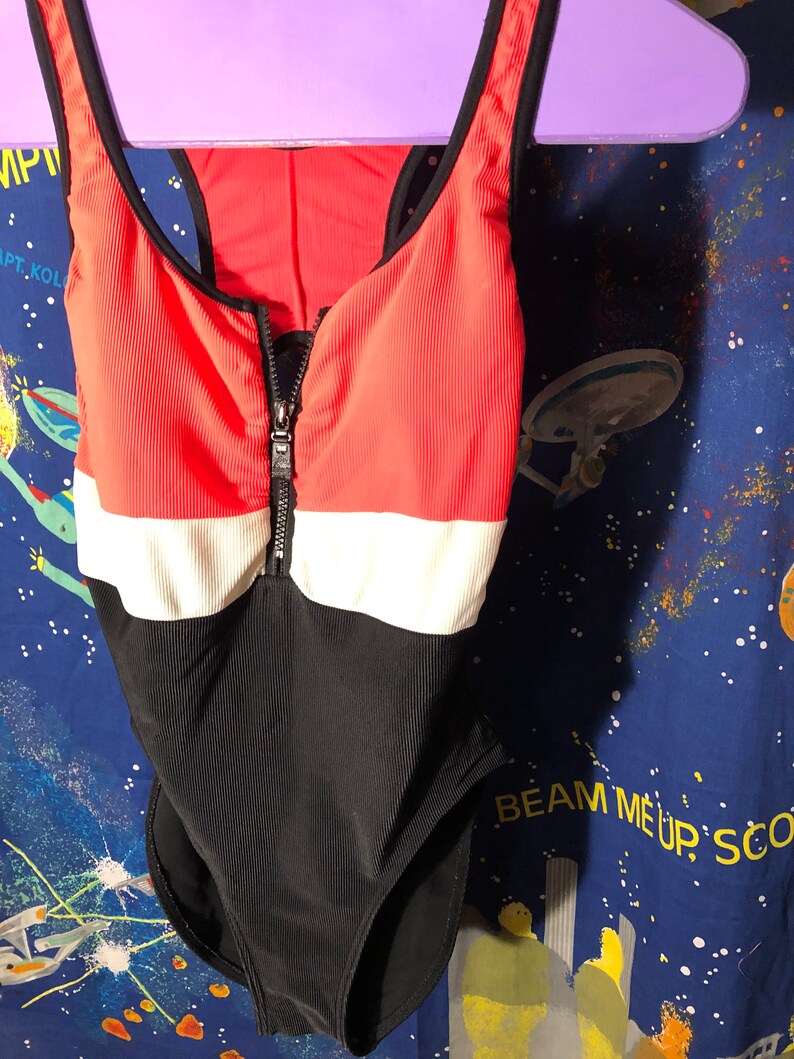 May include: A one-piece swimsuit with a color block design. The top section is coral, the middle is white, and the bottom is black. It has a zipper down the front and black straps. The swimsuit is hanging on a purple hanger.