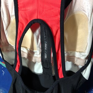 May include: A close-up of a black, red, and white swimsuit. The swimsuit has a ribbed texture on the black fabric and a zipper down the front. The straps and edges are black, and the center panel is red.