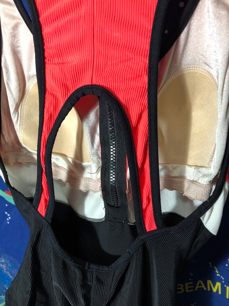 May include: A close-up of a black, red, and white swimsuit. The swimsuit has a ribbed texture on the black fabric and a zipper down the front. The straps and edges are black, and the center panel is red.