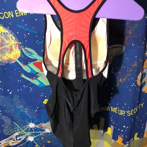 May include: A one-piece swimsuit with a black, white, and red color scheme. The back features a racerback design with a zipper closure. The swimsuit is hanging on a purple hanger.