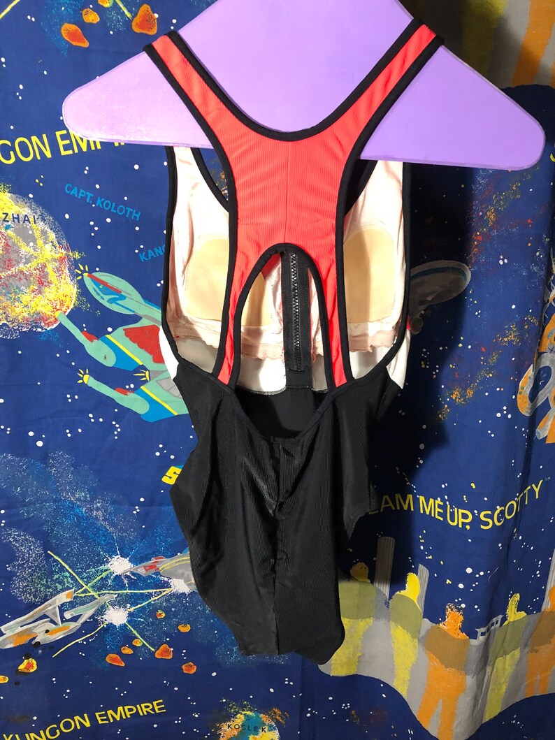 May include: A one-piece swimsuit with a black, white, and red color scheme. The back features a racerback design with a zipper closure. The swimsuit is hanging on a purple hanger.