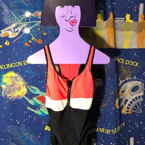 May include: A one-piece swimsuit with a color-block design in black, white, and coral. The swimsuit has a zipper down the front and is displayed on a purple hanger with a cartoon face. The background is a blue fabric with a space theme.