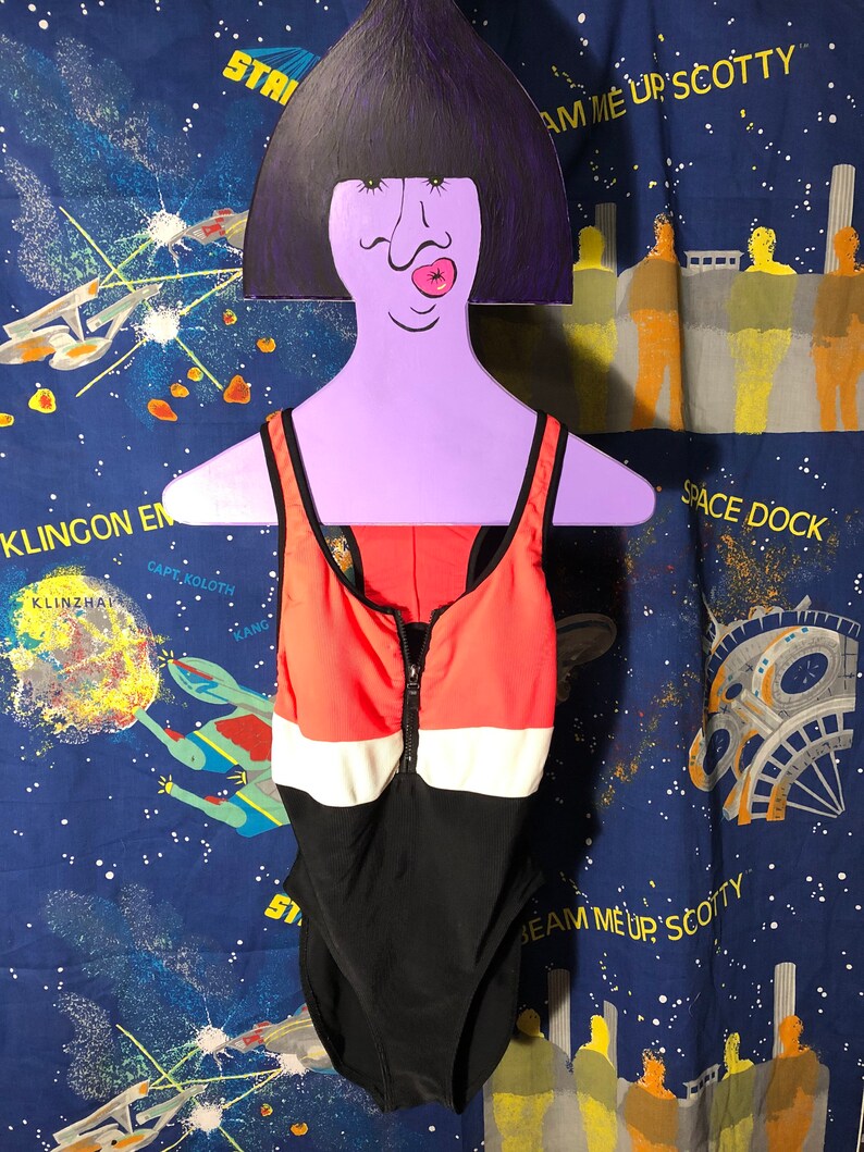 May include: A one-piece swimsuit with a color-block design in black, white, and coral. The swimsuit has a zipper down the front and is displayed on a purple hanger with a cartoon face. The background is a blue fabric with a space theme.