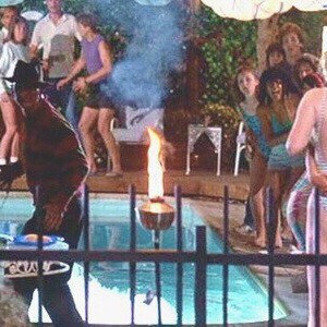 May include: A scene from a horror movie featuring a man in a striped sweater and hat, standing near a pool with a flaming torch. People in swimsuits and casual attire are in the background, suggesting a party setting. Smoke and a blue pool are also visible.