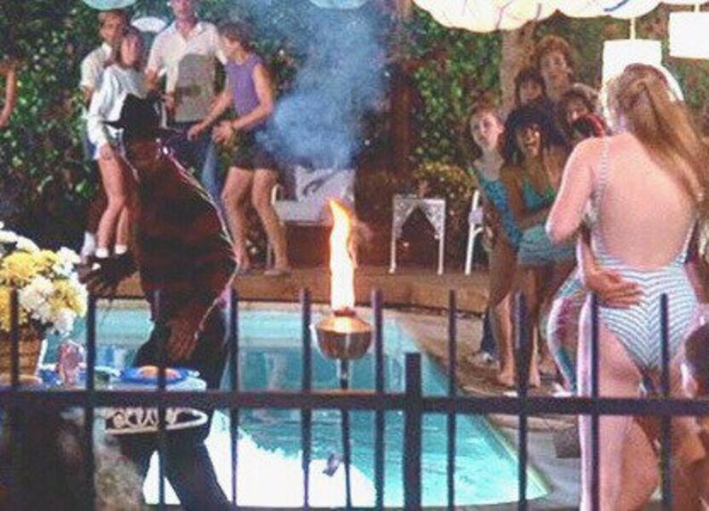 May include: A scene from a horror movie featuring a man in a striped sweater and hat, standing near a pool with a flaming torch. People in swimsuits and casual attire are in the background, suggesting a party setting. Smoke and a blue pool are also visible.