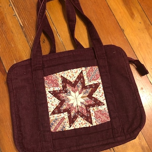 May include: A burgundy corduroy tote bag with a patchwork star design in shades of pink, brown, and white. The bag has two top handles and a zipper closure.