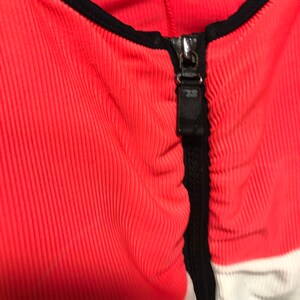 May include: Close-up of a coral-colored ribbed fabric garment with a black zipper and trim. The zipper is silver and has a black pull tab. White and black panels are visible on the sides.
