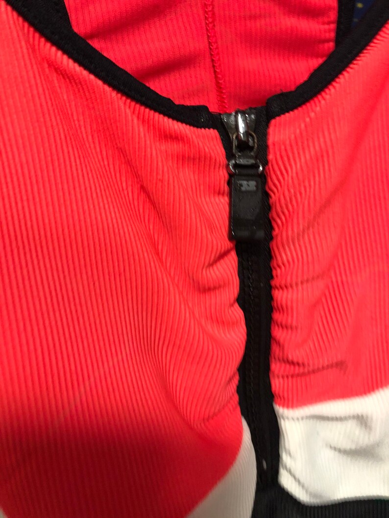 May include: Close-up of a coral-colored ribbed fabric garment with a black zipper and trim. The zipper is silver and has a black pull tab. White and black panels are visible on the sides.