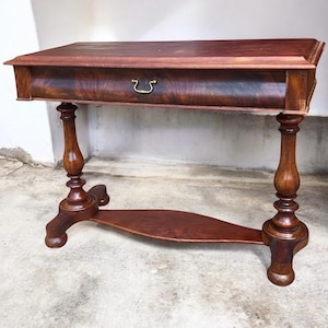 May include: A dark brown antique console table with a single drawer and ornate legs. The table features a curved lower shelf and a brass drawer pull. The wood grain is visible on the table's surface.