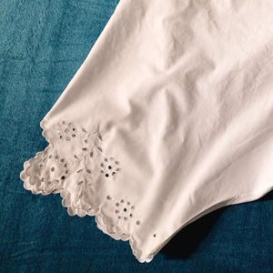 May include: White cotton bloomers with a scalloped, embroidered hem. The bloomers are laid flat on a blue surface, showcasing the details of the fabric and design. The garment appears to be vintage or antique.