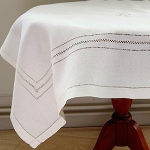 Puede incluir: Un mantel cuadrado blanco con un monograma bordado en el centro y bordes con dobladillo decorativo, colocado sobre una mesa de pedestal de madera marrón oscuro con tres patas curvas y pies en forma de garra en la base.