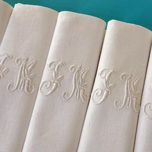 Puede incluir: Seis servilletas de lino blanco, cada una bordada con un monograma "K" en cursiva y detalles florales. Las servilletas están cuidadosamente dobladas y dispuestas sobre un fondo turquesa. Cada servilleta mide unos 40 cm de largo.