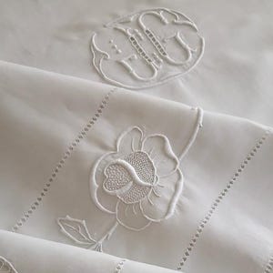 May include: White linen bedding with embroidered floral and monogram details. The bedding features a large embroidered flower, a monogram, and decorative stitching along the edges. The overall design is classic and elegant.